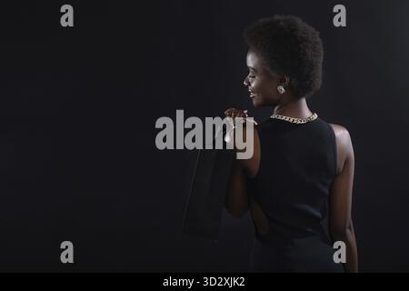 Femme afro-américaine vue arrière avec coiffure afro tient des sacs à provisions noirs. Soldes et réductions sur le marché et Black Friday concept Banque D'Images