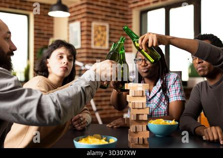 Groupe d'amis divers clinquant des bouteilles de bière, assis autour de la table avec des collations et des jeux. Les Caucasiens et les afro-américains apprécient un repaire intérieur dans une atmosphère chaleureuse et détendue. Banque D'Images