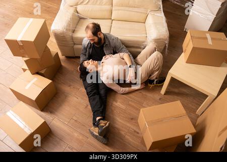 Homme barbu reposant sur le sol avec une belle femme, entouré de boîtes en carton, communiquant sur les meubles et les décorations de la maison. Moments heureux de relation des propriétaires caucasiens le jour du déménagement. Banque D'Images