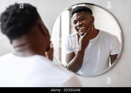 Homme noir souriant dans la salle de bain moderne tout en se préparant à se raser et prendre soin de sa peau Banque D'Images