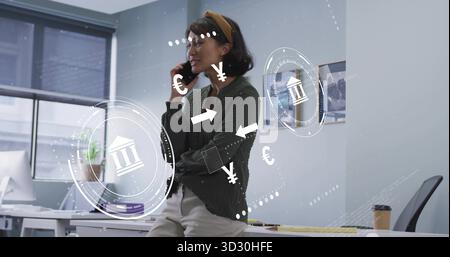 Femme asiatique penchée tenant smartphone à l'oreille et affichant des icônes bancaires numériques au bureau Banque D'Images