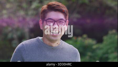 Rire homme asiatique mature portant un pull en tricot gris et des lunettes à bordure noire dans le jardin, lumière du jour Banque D'Images