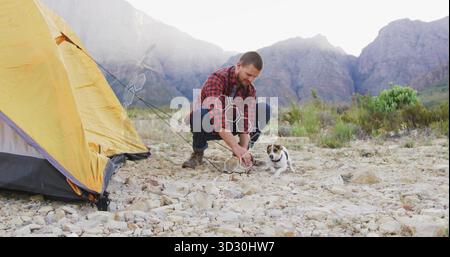Randonneur accroupi dans des bottes de réglage à carreaux au camping rocheux avec tente jaune et chien, espace de copie Banque D'Images