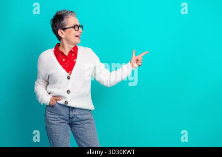 Femme mature amicale avec des cheveux gris courts et des lunettes portant un cardigan blanc et une chemise florale rouge pointent vers la droite contre une toile de fond sarcelle Banque D'Images