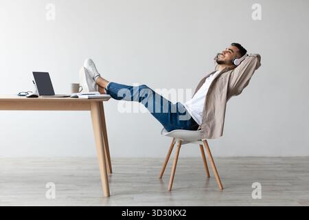 Homme relaxant appréciant la musique tout en étant assis au bureau avec les jambes sur la table dans l'espace de travail moderne Banque D'Images