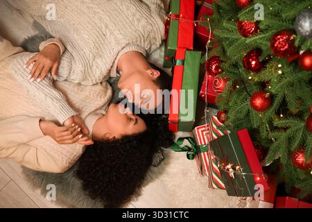 Heureux jeune couple dans des chandails chauds couchés sur le sol près de l'arbre de Noël et des cadeaux à la maison Banque D'Images