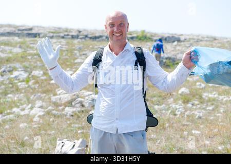 L'homme portant une chemise blanche et un short gris tient un sac bleu. Il sourit et fait signe Banque D'Images