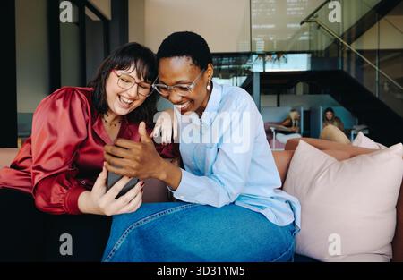 Deux jeunes femmes joyeuses assises sur un canapé, riant en regardant un smartphone, mettant en valeur le lien et le bonheur dans un cadre intérieur moderne. Banque D'Images