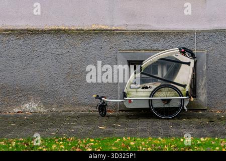 Une remorque de vélo se trouve stationnée à côté d'un mur texturé, présentant sa couverture transparente. De l'herbe verte et des feuilles tombées l'entourent, indiquant l'automne. Banque D'Images