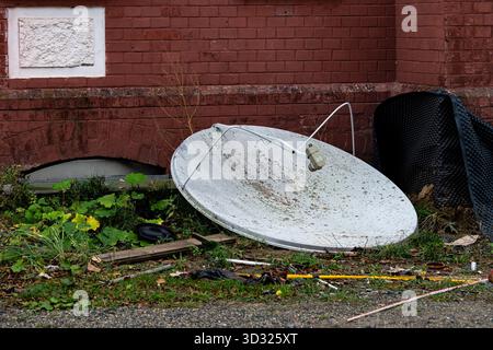 Une grande antenne parabolique cassée repose sur son côté à côté d'un bâtiment en briques. Autour, il est envahi par l'herbe et les débris, soulignant le St négligé Banque D'Images