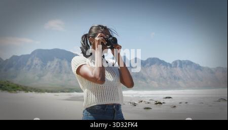 Photographe de cadrage portant un haut blanc et un Jean bleu, soulevant l'appareil photo reflex numérique au rivage avec des rochers Banque D'Images