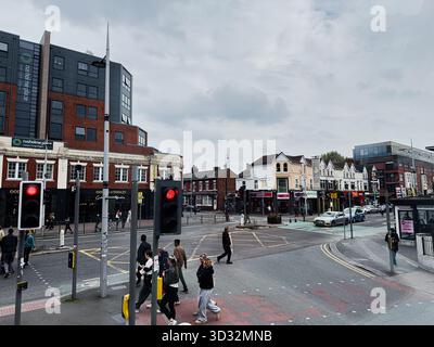 Manchester, Royaume-Uni - 29 septembre 2025 : les gens traversent à un carrefour animé alors que les voitures et les bus passent au milieu des magasins et des appartements urbains. Banque D'Images