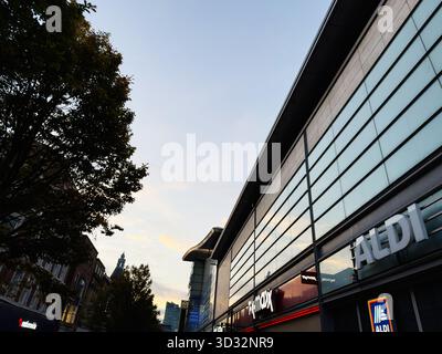 Manchester, Royaume-Uni - 29 septembre 2025 : la lumière du coucher du soleil se reflète sur les vitrines le long d'une rue de Manchester à côté des magasins ALDI à proximité. Banque D'Images