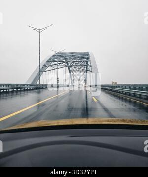 Point de vue : conduite le long d'un pont le jour de pluie. Vue en perspective de l'intérieur de la voiture : la route devant, arche métallique du pont. Banque D'Images