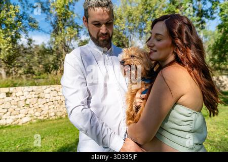 Heureux couple embrassant leur petit chien dans un parc ensoleillé créant des liens familiaux affectueux Banque D'Images
