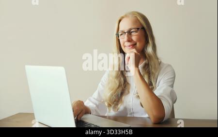 Heureuse femme mature confiante travaillant sur ordinateur portable portant des lunettes, assis au bureau au bureau ou à la maison || modèle libéré Banque D'Images