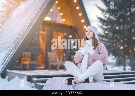 Agréable moment d'hiver d'une jeune femme profitant du plein air enneigé devant une charmante cabane au coucher du soleil. Banque D'Images