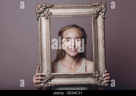 Jeune femme millénial cheveux blonds tient image dorée cadre dans les mains visage portrait Banque D'Images