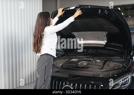 Jeune femme debout devant la voiture avec le capot ouvert regardant sous le capot de la voiture Banque D'Images