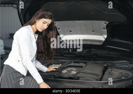 Jeune femme debout devant la voiture avec le capot ouvert regardant sous un capot de voiture Banque D'Images