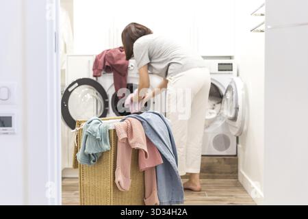 Jeune femme chargeant la machine à laver et le panier plein de vêtements sales dans la buanderie Banque D'Images