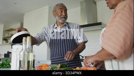 Couple senior dans des tabliers soulevant le couvercle du cuiseur à riz tranchant des carottes sur une planche à découper à l'îlot de cuisine Banque D'Images