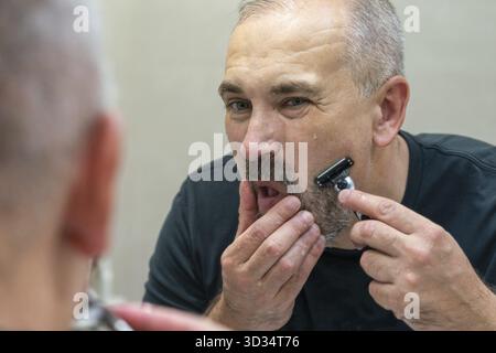 Charmant homme d'âge moyen rasant sa barbe et sa moustache devant le miroir pour paraître plus jeune. Homme mûr aux cheveux gris restylant sa barbe lui-même Banque D'Images