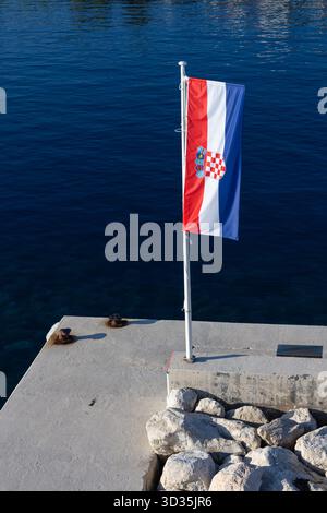 Un drapeau croate agite sur un mât blanc à côté d'une jetée en béton, surplombant les eaux bleu profond de l'Adriatique. La scène transmet le calme maritime, scen côtier Banque D'Images