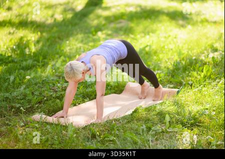 La femme d'âge moyen exerce et fait des exercices de renforcement de base. Femme âgée pratique le fitness et le yoga à l'extérieur dans le parc le jour ensoleillé, la disposition f Banque D'Images