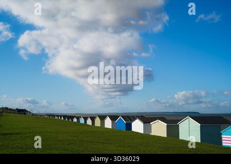 Une rangée de cabanes de plage en bois colorées à Tankerton, Whitstable. Un grand nuage blanc domine le ciel bleu. L'île de Sheppey est à l'horizon. Banque D'Images