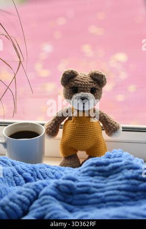 Ours en peluche tricoté en combinaison jaune avec tasse à café sur couverture bleue, scène à la maison confortable. Banque D'Images