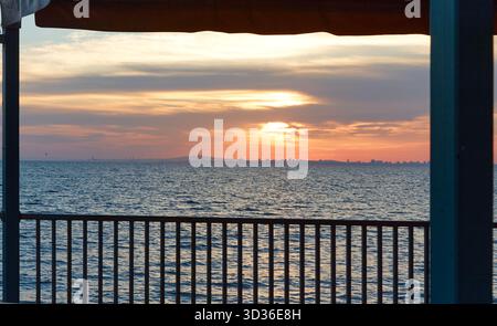 Vue tranquille sur le coucher du soleil sur les eaux de mer paisibles, encadrée par une balustrade en bois, avec des teintes vibrantes d'orange, rose et violet dans le ciel, reflétant magnifiquement Banque D'Images