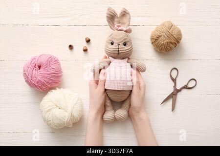 Mains féminines avec jouet lapin tricoté, fil et ciseaux sur fond de bois blanc Banque D'Images