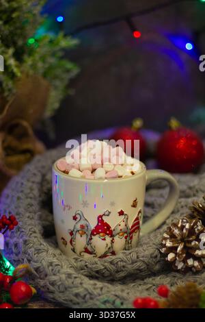 Une tasse confortable remplie de guimauves roses et blanches est entourée de pommes de pin, d'ornements festifs et de lumières scintillantes, capturant des vacances festives Banque D'Images