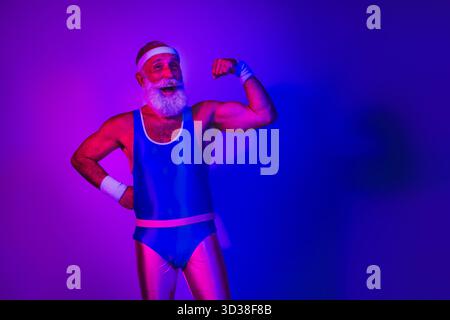 Homme âgé énergique en combinaison rétro bleu fléchissant bras sous des lumières bleues violettes néon dans une scène de fitness ludique Banque D'Images
