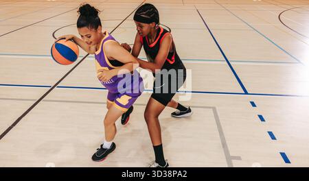 Deux jeunes femmes participent activement à un match de basket-ball sur un terrain de sport couvert. Ils font preuve de travail d'équipe et de compétence dans un cadre dynamique et compétitif Banque D'Images