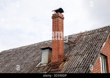 Gros plan d'un toit en pente avec des tôles ondulées altérées, une grande cheminée en briques rouges, un conduit métallique et une échelle d'accès pour l'entretien. Banque D'Images