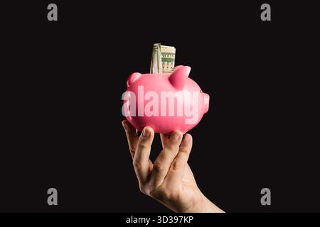La main d'un homme tient une tirelire rose avec un billet d'un dollar américain sortant de la fente, isolé sur un fond noir Uni. Banque D'Images