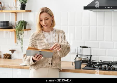 Femme blonde profite d'un moment de lecture paisible tout en étant habillée confortablement dans une cuisine moderne. Banque D'Images