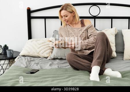 Une femme vêtue de vêtements confortables se détend sur son lit, lisant un livre à la maison. Banque D'Images