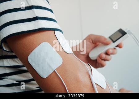 un homme utilisant un stimulateur musculaire sur son bras, avec des électrodes attachées à la peau pendant une séance de rééducation par électrothérapie Banque D'Images