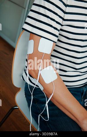 un homme portant une chemise rayée utilisant un appareil de stimulation musculaire sur son bras, assis à l'intérieur pendant un traitement de physiothérapie Banque D'Images