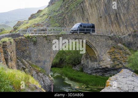 Un van noir conduit sur un vieux pont de pierre, entouré par un paysage rocheux pittoresque et fluvial, camping-car, sur le pont Melik Tangi, derrière Banque D'Images