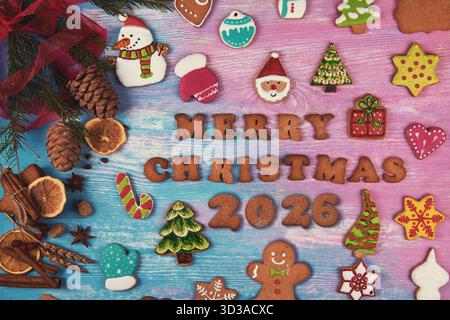 Des biscuits de Noël colorés en forme de bonhommes de neige, d'arbres, d'étoiles et de pain d'épice entourent le texte Joyeux Noël 2026 sur un fond en bois. Banque D'Images