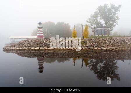 Une vue horizontale atmosphérique d'un phare décoratif rouge et blanc et d'une petite structure de parc reflétée dans l'eau calme sur un autu dense et brumeux Banque D'Images