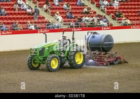Calgary, Alberta, Canada. 12 juillet 2025. Un tracteur John Deere vert arrose le sol de terre de l'arène lors d'un événement de rodéo, avec des spectateurs visibles dans la Re Banque D'Images