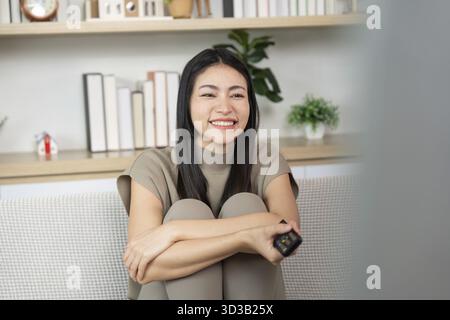 Une jeune femme joyeuse s'assoit confortablement tout en regardant la télévision, rayonnant de bonheur dans un salon confortable. Concept de détente et de divertissement Banque D'Images
