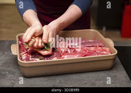 Nourriture, gros plan d'un chef pressant du romarin et de la sauge sur des coupes de bœuf cru dans un plat allant au four, préparant pour la marinade ou la cuisson Banque D'Images