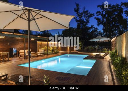 Piscine arrière avec terrasse en bois et parasol élégant sous le ciel du soir. Salon extérieur moderne avec ambiance sereine et éclairage élégant Banque D'Images