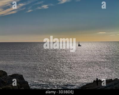 Un seul voilier glisse à travers l'Atlantique scintillant au large de la côte bretonne en France, rétroéclairé par une lumière chaude de fin d'après-midi avec le rivage rocheux en vue. Banque D'Images